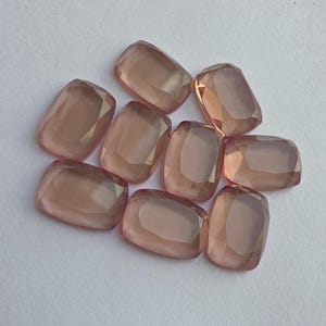 May include: A collection of nine faceted, rectangular gemstones in a soft peach color. The gems have rounded corners and are arranged on a white surface. Each stone reflects light, showcasing its cut and clarity.