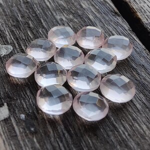 May include: A group of 10 pale pink rose quartz gemstones, each with a faceted, rounded shape. The stones are arranged in a loose cluster on a wooden surface.