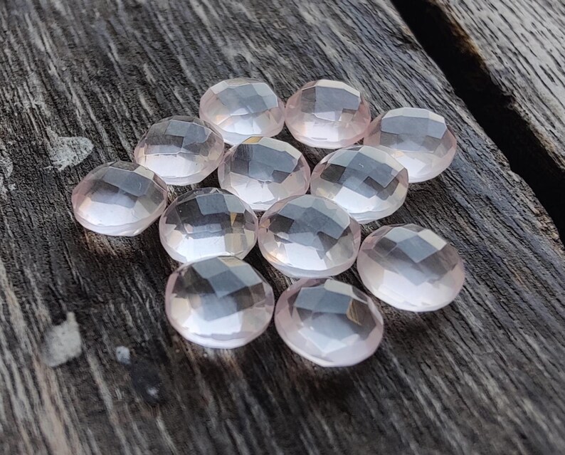 May include: A group of 10 pale pink rose quartz gemstones, each with a faceted, rounded shape. The stones are arranged in a loose cluster on a wooden surface.