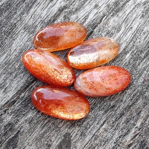 K&ouml;nnte beinhalten: F&uuml;nf glatte, ovalf&ouml;rmige Sunstone-Steine mit schimmerndem, irisierendem Finish. Die Steine sind in Orange- und Rott&ouml;nen gehalten.