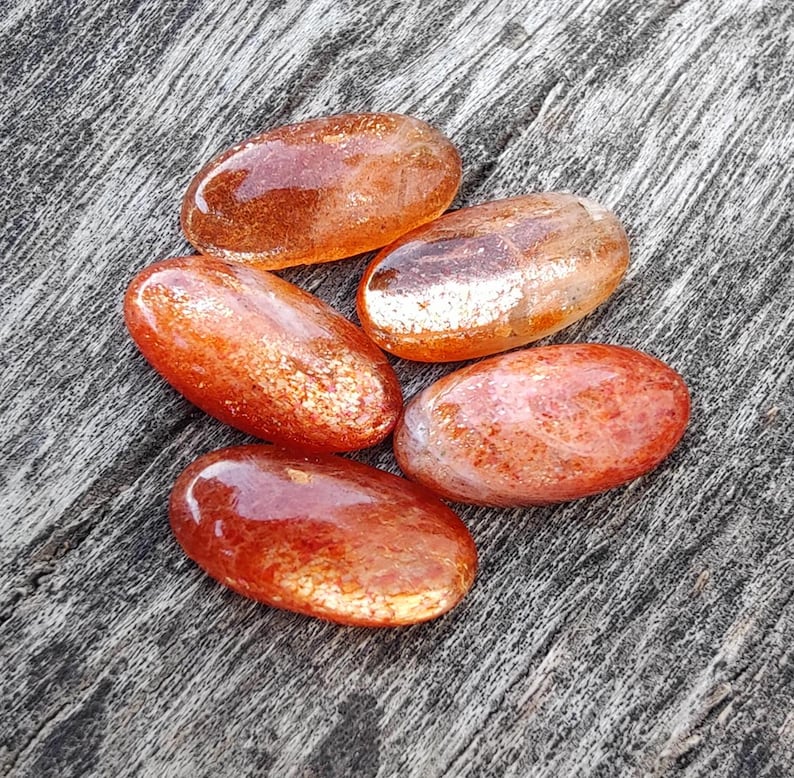 K&ouml;nnte beinhalten: F&uuml;nf glatte, ovalf&ouml;rmige Sunstone-Steine mit schimmerndem, irisierendem Finish. Die Steine sind in Orange- und Rott&ouml;nen gehalten.