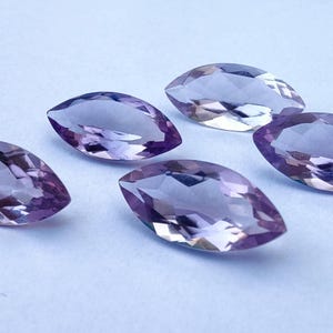 Puede incluir: Cinco gemas de talla marquesa con superficie facetada. Las gemas son de color púrpura claro con un ligero degradado, dispuestas sobre una superficie blanca. Las gemas son probablemente amatistas.
