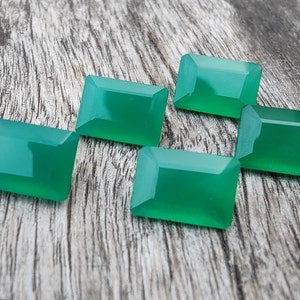 May include: Five green gemstone cabochons, cut in a rectangular shape, are arranged on a wooden surface. The gemstones are a vibrant green color and have a smooth, polished finish.