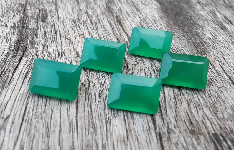 May include: Five green gemstone cabochons, cut in a rectangular shape, are arranged on a wooden surface. The gemstones are a vibrant green color and have a smooth, polished finish.