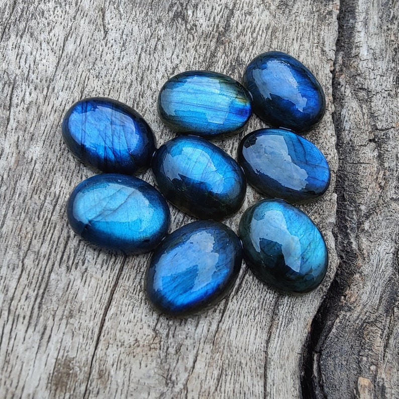 May include: A group of eight oval-shaped labradorite gemstones in shades of blue and green. The stones are arranged in a cluster on a wooden surface.