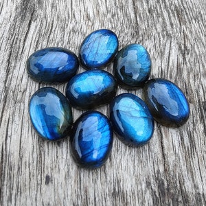 May include: A collection of eight oval-shaped labradorite gemstones, each with a unique blue and green iridescent sheen. The stones are arranged in a cluster on a wooden surface.