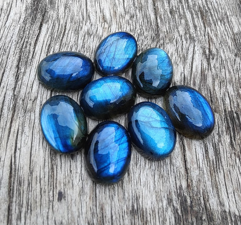 May include: A collection of eight oval-shaped labradorite gemstones, each with a unique blue and green iridescent sheen. The stones are arranged in a cluster on a wooden surface.