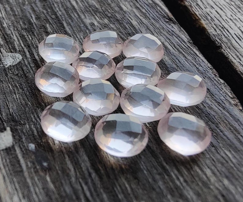 May include: A collection of 12 faceted rose quartz stones, each with a light pink hue and a translucent appearance. The stones are arranged in a loose cluster on a wooden surface.
