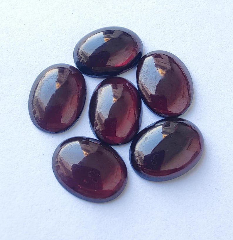 Puede incluir: Seis piedras preciosas ovaladas de color rojo oscuro con un acabado brillante. Las piedras preciosas est&aacute;n dispuestas en un c&iacute;rculo sobre un fondo blanco.