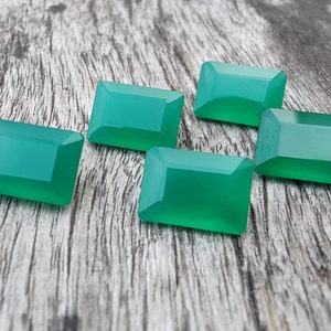 May include: Six emerald green rectangular gemstones on a wooden surface.