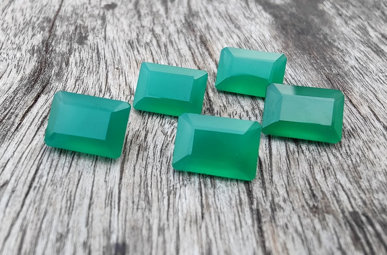 May include: Six emerald green rectangular gemstones on a wooden surface.