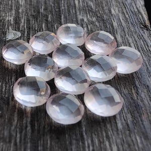 May include: A collection of twelve faceted, oval-shaped, pale pink gemstones. The stones are arranged in a cluster on a wooden surface.