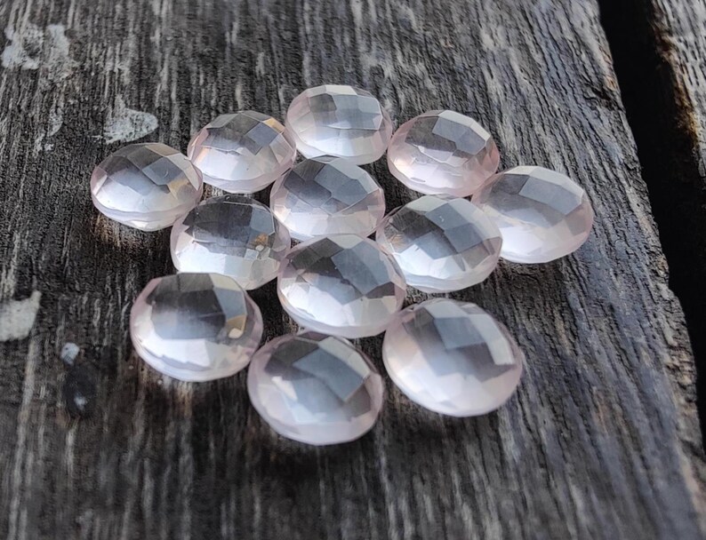 May include: A collection of twelve faceted, oval-shaped, pale pink gemstones. The stones are arranged in a cluster on a wooden surface.