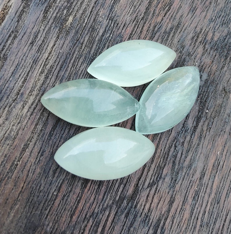 May include: Four light green, translucent, oval-shaped stones on a dark brown wooden surface.