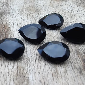 May include: Five black faceted teardrop-shaped gemstones on a wooden surface.