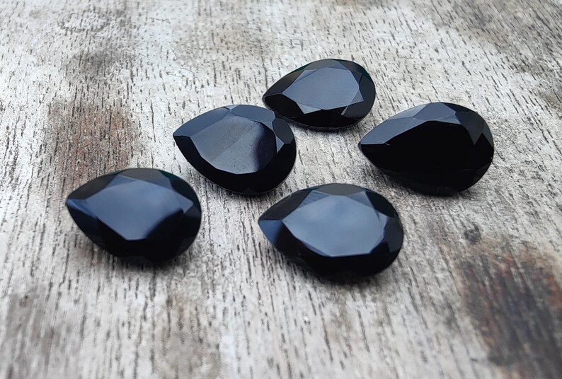 May include: Five black faceted teardrop-shaped gemstones on a wooden surface.