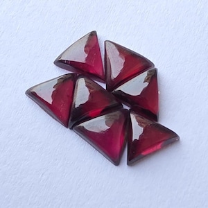 May include: A cluster of triangular, deep red gemstones. The polished stones are arranged on a white surface, showcasing their glossy, reflective surfaces. These gems are likely for jewellery making or crafting.