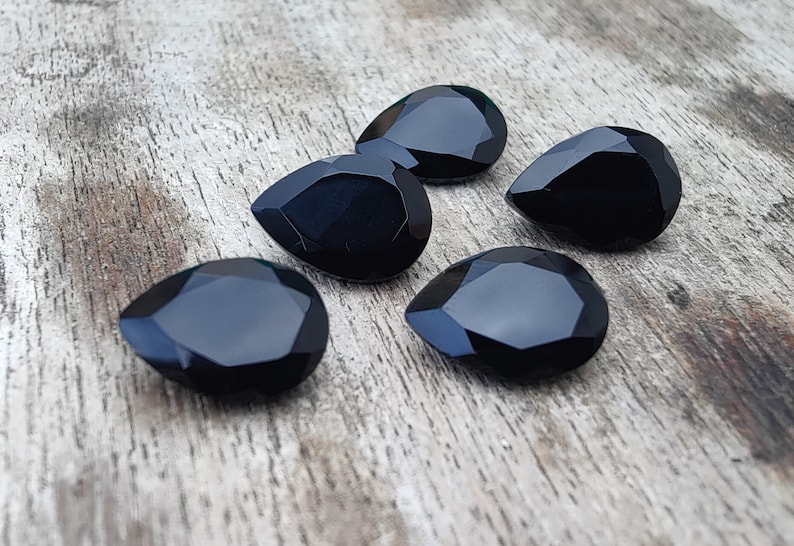 May include: Five black teardrop-shaped faceted gemstones on a wooden surface.