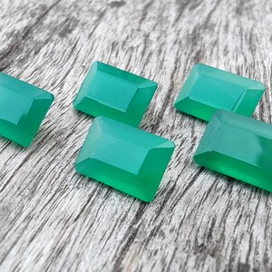 May include: Six emerald green gemstone cabochons, cut in a rectangular shape, are arranged on a wooden surface.