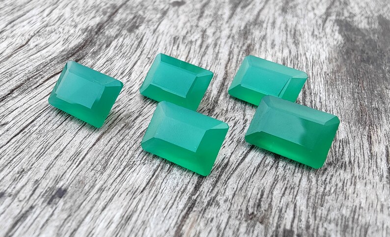 May include: Six emerald green gemstone cabochons, cut in a rectangular shape, are arranged on a wooden surface.