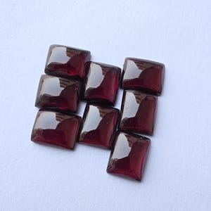 Puede incluir: Nueve piedras preciosas cuadradas pulidas, de color burdeos oscuro, están dispuestas sobre una superficie blanca. Cada piedra tiene esquinas redondeadas y una superficie reflectante, que capta la luz. Las gemas son probablemente para la fabricación de joyas.