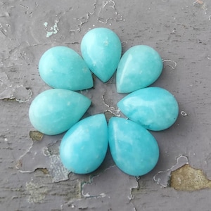 Pode incluir: Sete pedras preciosas polidas em forma de gota, em tom turquesa claro, dispostas em padrão circular. As pedras têm uma superfície lisa e brilhante, sobre um fundo cinza desgastado.