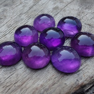 May include: Eight round, purple amethyst cabochons, polished and smooth, arranged in a cluster on a wooden surface.