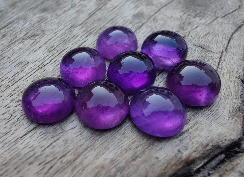 May include: Eight round, purple amethyst cabochons, polished and smooth, arranged in a cluster on a wooden surface.