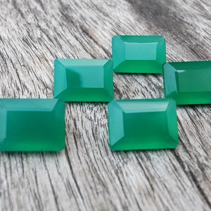May include: Six emerald green rectangular gemstones on a wooden surface.