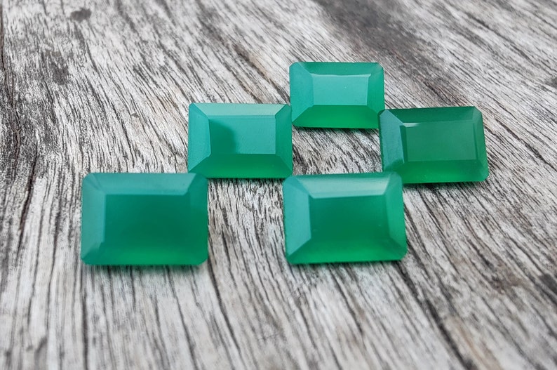 May include: Six emerald green rectangular gemstones on a wooden surface.