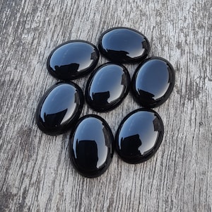 Peut inclure: Sept pierres précieuses noires polies, de forme ovale, disposées sur une surface en bois vieilli. Les pierres ont une finition très brillante, reflétant la lumière. Les pierres sont probablement utilisées pour la joaillerie ou comme éléments décoratifs.