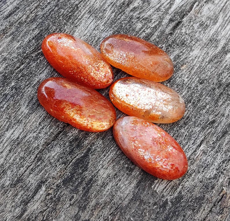 K&ouml;nnte beinhalten: F&uuml;nf ovale Sunstone-Steine mit schimmerndem, irisierendem Finish. Die Steine sind eine Mischung aus Orange und Rot mit einem metallischen Schimmer.