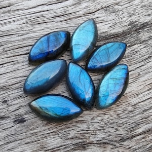 Op de afbeelding: Zeven iriserende blauwe labradorit edelsteen cabochons in een traanvorm, gerangschikt in een cluster op een houten oppervlak.