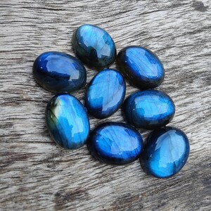 May include: Eight oval-shaped labradorite gemstones in shades of blue and green. The stones are arranged in a cluster on a wooden surface.