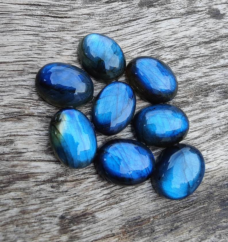 May include: Eight oval-shaped labradorite gemstones in shades of blue and green. The stones are arranged in a cluster on a wooden surface.