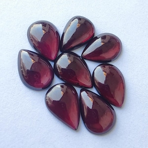 Puede incluir: Ocho gemas pulidas en forma de lágrima, en varios tonos de rojo oscuro y burdeos. Las piedras tienen una superficie lisa y reflectante, dispuestas sobre un fondo claro. Estas gemas son adecuadas para la fabricación de joyas.