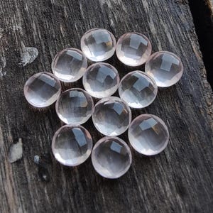 May include: A collection of twelve faceted rose quartz gemstones, each with a light pink hue and a smooth, polished finish. The stones are arranged in a circular pattern on a wooden surface.