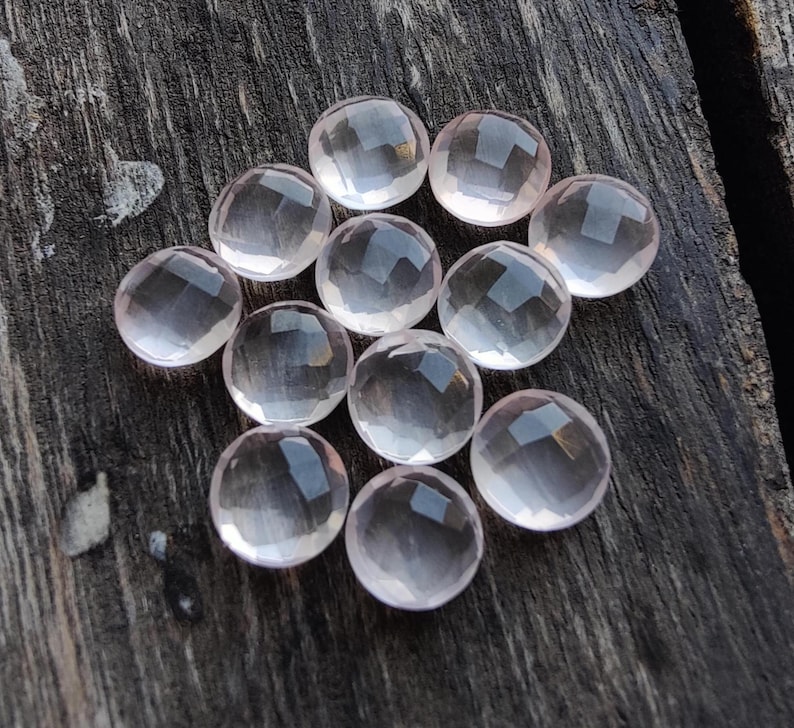 May include: A collection of twelve faceted rose quartz gemstones, each with a light pink hue and a smooth, polished finish. The stones are arranged in a circular pattern on a wooden surface.