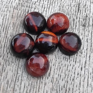Puede incluir: Seis cabujones de ojo de tigre rojo, cada uno con un patrón diferente de rayas marrones y doradas. Los cabujones son redondos y lisos, y están dispuestos en un grupo sobre una superficie de madera.