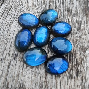 May include: Eight oval-shaped labradorite gemstones with iridescent blue and green hues. The stones are arranged in a cluster on a wooden surface.