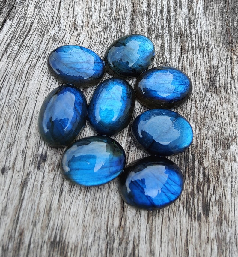 May include: Eight oval-shaped labradorite gemstones with iridescent blue and green hues. The stones are arranged in a cluster on a wooden surface.