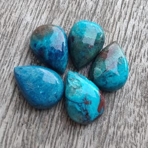 Pode incluir: Cinco pedras preciosas em forma de gota em tons de verde-azulado e turquesa, com inclusões castanhas e vermelhas. As pedras polidas estão dispostas sobre uma superfície de madeira cinzenta desgastada. As pedras preciosas têm uma superfície brilhante e reflectora.