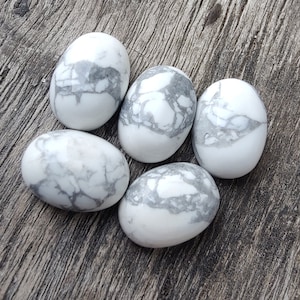 Puede incluir: Cinco piedras de howlita blancas, ovaladas, con vetas grises y pulidas. Se disponen sobre una superficie de madera envejecida, mostrando sus patrones naturales. Estas piedras se usan a menudo para la meditación y la curación.