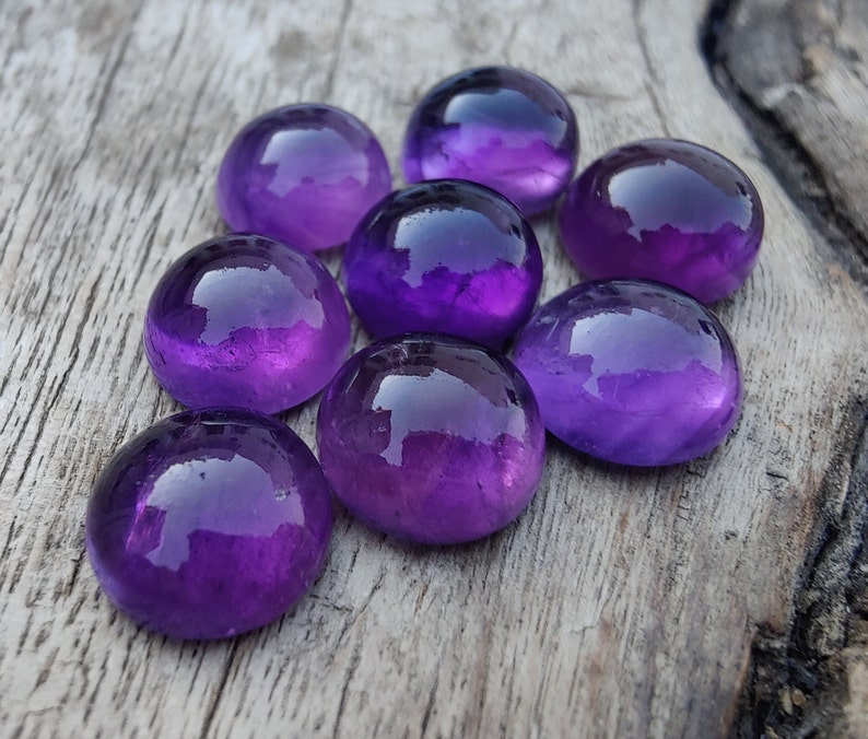 May include: Eight smooth, round, purple amethyst gemstones on a grey wooden surface.