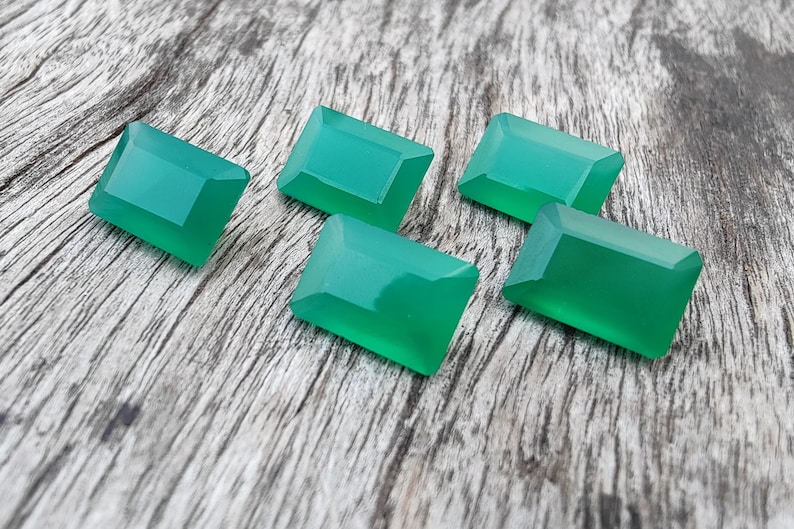 May include: Five emerald green gemstone cabochons in a rectangular shape, displayed on a wooden surface.
