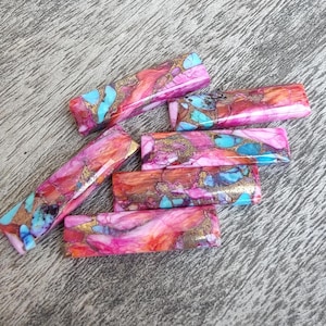 Peut inclure: Des perles de pierres précieuses rectangulaires dans les tons de rose, de rouge, d'orange et de turquoise avec des accents dorés. Les perles sont disposées sur une surface en bois. Ces perles pourraient être utilisées pour la fabrication de bijoux.