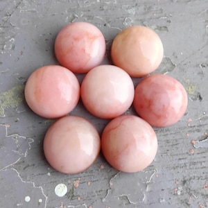May include: Seven pink opal cabochons, each with a slightly different pattern of white and pink swirls. The cabochons are smooth and polished, and they are arranged in a circle on a gray background.