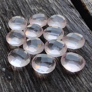 May include: A collection of twelve pale pink, faceted, oval-shaped gemstones. The stones are arranged in a cluster on a wooden surface.