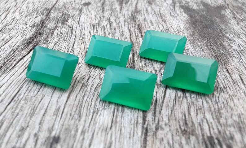May include: Five green gemstone cabochons in a rectangular shape, with a smooth, polished surface. The stones are arranged on a wooden surface.