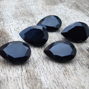 May include: Five black teardrop-shaped faceted gemstones on a wooden surface.